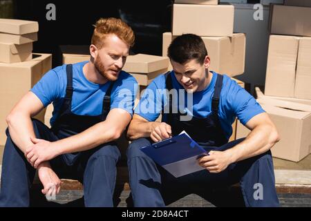 Movers looking at clipboard while sitting near cardboard boxes in warehouse Stock Photo