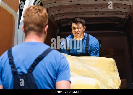 Selective focus of loader in uniform holding couch in stretch wrap with colleague near truck outdoors Stock Photo