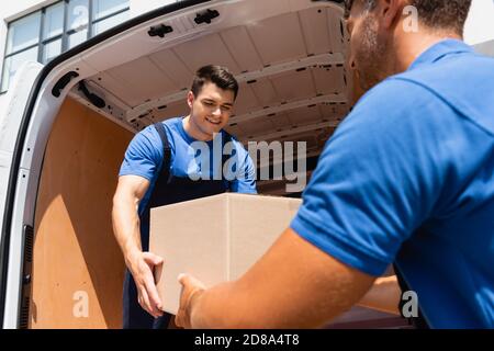 Selective focus of loader holding cardboard box with colleague near truck on urban street Stock Photo