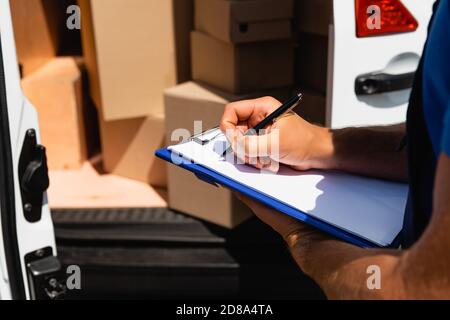 Cropped view of loader writing on clipboard near carton boxes in truck outdoors Stock Photo