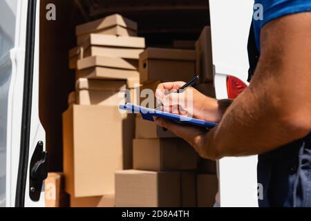 Cropped view of loader writing on clipboard beside cardboard boxes in truck outdoors Stock Photo