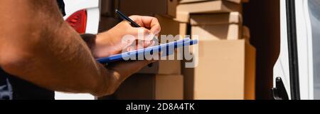 Panoramic crop of loader writing on clipboard with cardboard boxes in truck at background Stock Photo