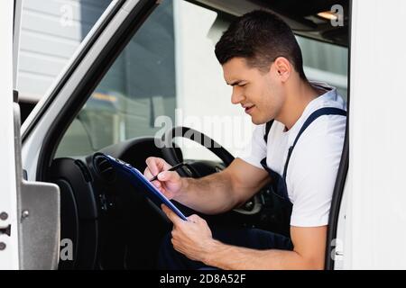 Selective focus of concentrated loader writing on clipboard while sitting on driver seat in truck Stock Photo