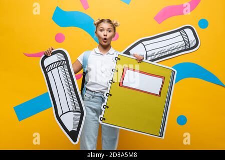 Surprised schoolgirl holding paper pencil and notebook on yellow background Stock Photo