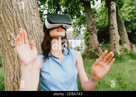 selective focus of young woman in virtual reality headset gesturing near green trees Stock Photo