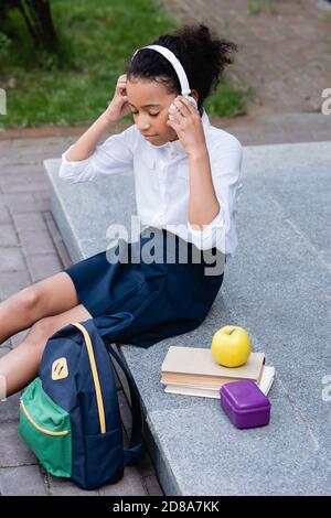 african american schoolgirl listening music in headphones near backpack, books and lunch outdoors Stock Photo
