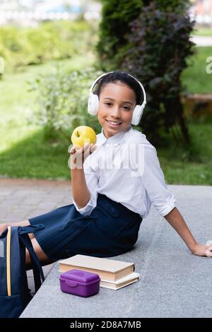 smiling african american schoolgirl in headphones holding apple near backpack, books and lunch box outdoors Stock Photo