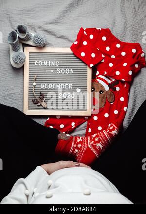 Pregnant woman sitting with a coming soon December 2020 Baby announcement sign. Coming soon Christmas concept. Pregnancy belly. Stock Photo