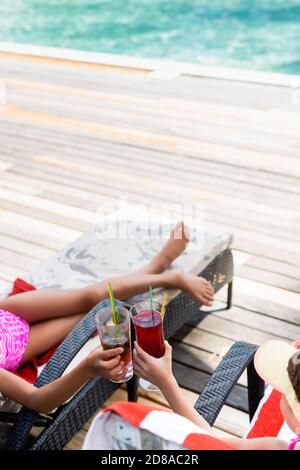 high angle view of girls sitting in sunbeds and clinking glasses with fresh cocktails Stock Photo