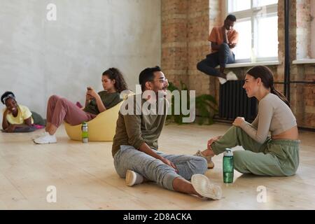 Young couple of dancers sitting on the floor and talking to each other in dance studio with other dancers in the background Stock Photo