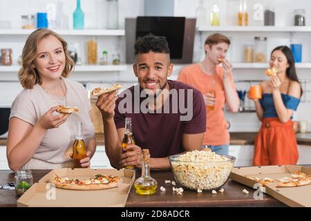 pleased multiethnic friends looking at camera while eating pizza during party Stock Photo