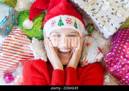 Christmas time! Joyful excited young woman lying surrounded by many gifts wearing a red Santa hat. Stock Photo