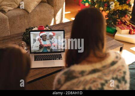 Rear view of woman and son having a videocall with senior couple in santa hats opening gift box on l