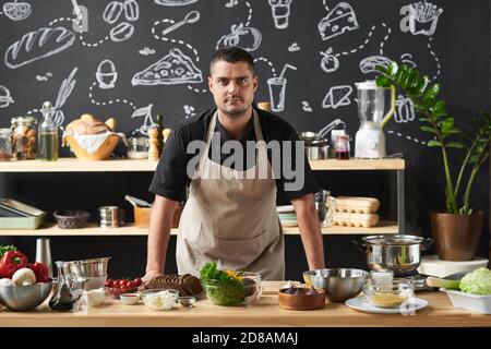 Portrait of confident cook in apron looking at camera while standing near the table with food in the kitchen Stock Photo
