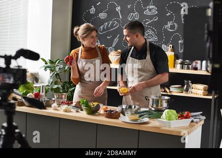 Happy young couple in aprons standing in the kitchen and discussing the new recipe together they shooting the blog Stock Photo
