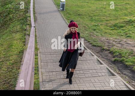 A joyful girl walks up the stairs in a burgundy palette and biret, with a beautiful smile in black clothes, in the fall against a background of blue Stock Photo