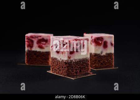 Front view of three delicious fresh pieces of cake made with brown biscuit and mousse pink layer with red jelly cherries. Tasty sweet dessert isolated on black background at restaurant. Stock Photo
