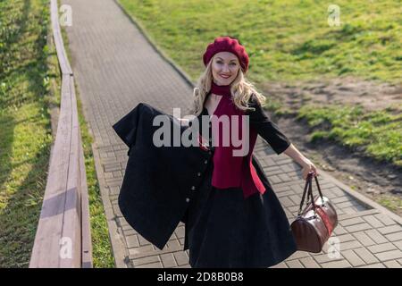 A joyful woman walks up the stairs in burgundy palla and biret, with beautiful eyes in black clothes, in the fall against the background of a pond Stock Photo