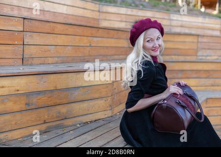 Cheerful sits on a summer theater bench, wooden in a burgundy coat and biret, an adult smiles at the camera, in the fall against the background of a Stock Photo