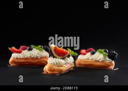 Front view of fresh dessert in row isolated on black background. Baked homemade eclairs filled with cream, white topping and slices of figs, blackberries, raspberries and blueberries. Stock Photo