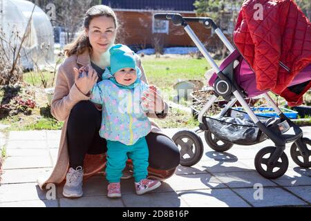 Mother holding her cheerful baby boy at house courtyard, toddler standing next to baby buggy Stock Photo