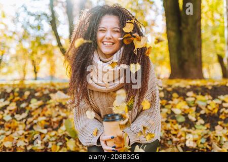 Positive afro hair woman with beautiful smile wearing knitted sweater and scarf throw yellow autumn leaves in park on sunny day. Stock Photo