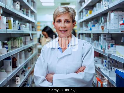 Smiling pharmacist with cross arms standing in medication isle wearing lab coat in pharmacy Stock Photo