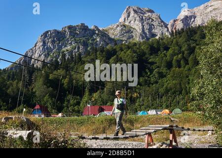 young woman hiker walks on a hinged bridge across the river against the background of a campground in a mountain valley Stock Photo