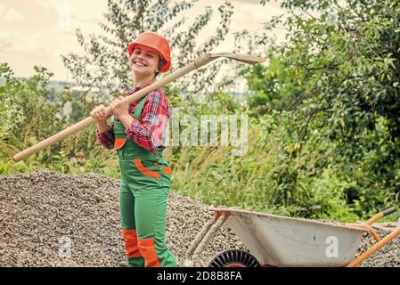 teen girl takes out rubble from wheelbarrow. kid with shovel loading crushed stones. Laying the foundation, building project. girl with wheelbarrow of rubble. kid working on construction site. Stock Photo