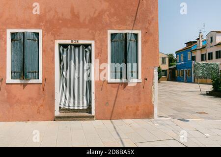 Brown wall house in Burano, with two green windows and a striped curtain and view over a small empty square Stock Photo