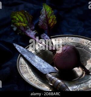 Whole fresh beet with foliage placed on vintage plate with kitchen knife against black background Stock Photo