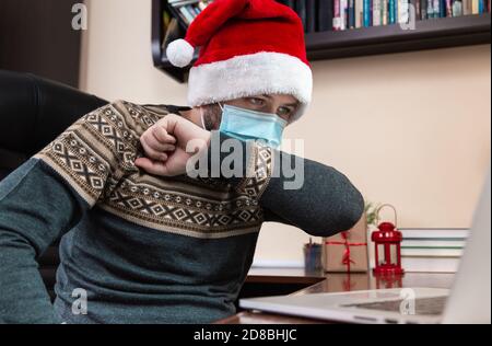 Christmas disease. Young man in santa claus hat and face mask coughs in the elbow and talks using laptop for video call friends and childrens. Christm Stock Photo