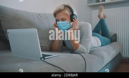 Young blond caucasian woman lying on the couch and working on laptop with medical mask. Work and education during quarantine. High quality photo Stock Photo