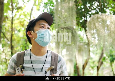 Young adult Asian travel backpacker wearing a face mask hiking in the forest. Millenial man wearing a cap walking in the nature during Covid-19 or Corona Virus pandemic. Stock Photo