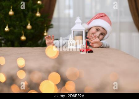 Toy red car carries a bag with gifts. A child peeking out from behind the house waves with two hands, greeting. Stock Photo