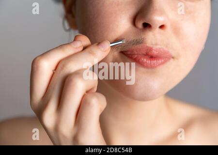 A young woman plucks her hair over her upper lip with tweezers. The concept of getting rid of unwanted facial hair. Stock Photo