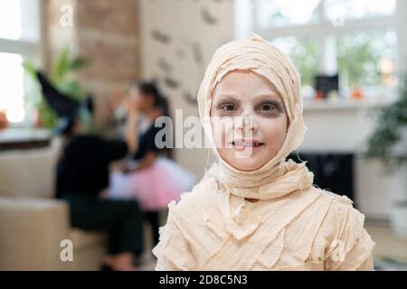 Portrait of smiling boy with white face wrapped up into bandages like mummy standing in room decorated for Halloween party Stock Photo