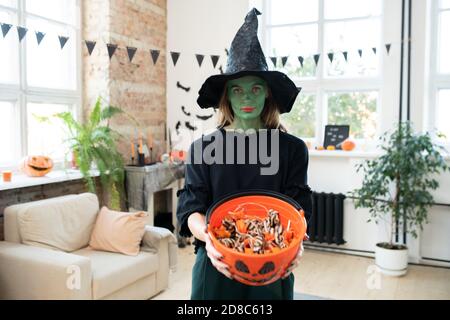 Portrait of serious young woman with green face wearing witches costume holding Halloween bucket of treats in decorated living room Stock Photo