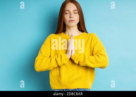 Cheerful caucasian freckled redhead woman making praying gesture, keeping palms together, asking husband to buy her new handbag isolated over blue bac Stock Photo