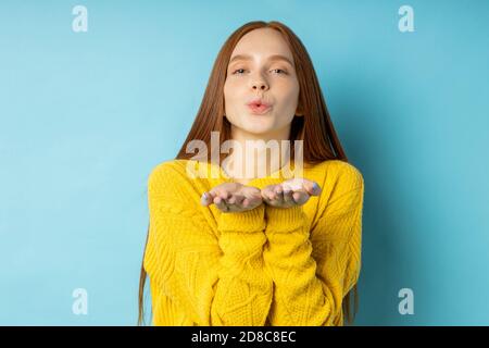 Lovely redhed caucasian girl in yellow sweater with no makeup blowing air kiss, stretching palms, expressing love and affection isolated over blue stu Stock Photo