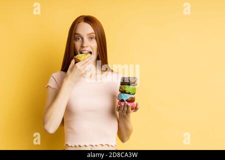 Joyful caucasian redhead young woman eating delicious pastry, holding sweet glazed doughnuts, enjoying tasty dessert isolated over yellow background. Stock Photo