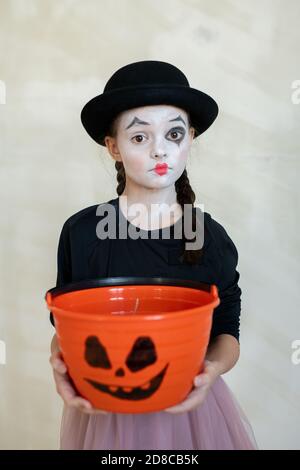 Portrait of serious shocked mime girl in black hat holding orange treat bucket with scary face image against isolated background, Halloween concept Stock Photo