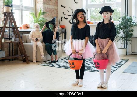 Serious multi-ethnic mimes girls in pink tutu skirts and black hats standing with trick-or-treating buckets in living room with Halloween decorations Stock Photo