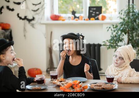 Group of happy kids in Halloween costumes sitting at table in decorated living room and eating cookies and candies at tea party Stock Photo