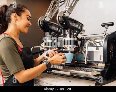Female barista using a coffee machine. Smiling woman pouring hot coffee into cup at cafe. Stock Photo