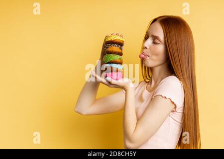 caucasian woman with red hair, closed eyes, licking her lips with tongue, holding in hands delicious colorful donuts isolated over yellow background. Stock Photo