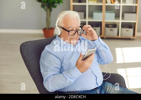 Aged man in shirt, glasses and headphones sitting in armchair and listening to music on smartphone Stock Photo