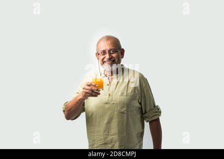 Indian asian senior or old man drinking fresh orange juice in a glass while standing against white background Stock Photo
