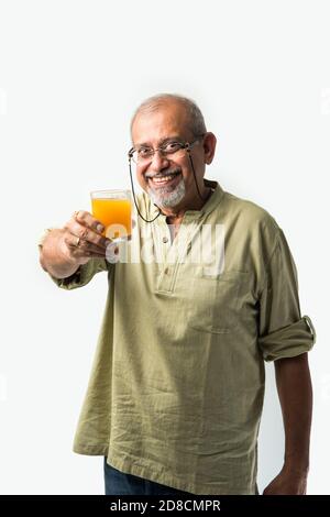 Indian asian senior or old man drinking fresh orange juice in a glass while standing against white background Stock Photo