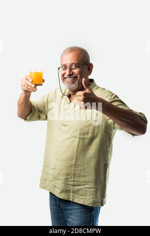 Indian asian senior or old man drinking fresh orange juice in a glass while standing against white background Stock Photo